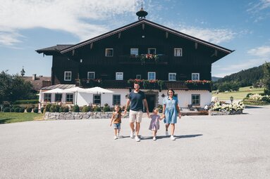 Traditionelles Holzhaus mit Blumen und Garten, davor eine Familie mit zwei Kindern, die durch die Sonne spazieren. Eine idyllische Atmosphäre in einer ländlichen Umgebung. | © Urlaub am Bauernhof / Daniel Gollner